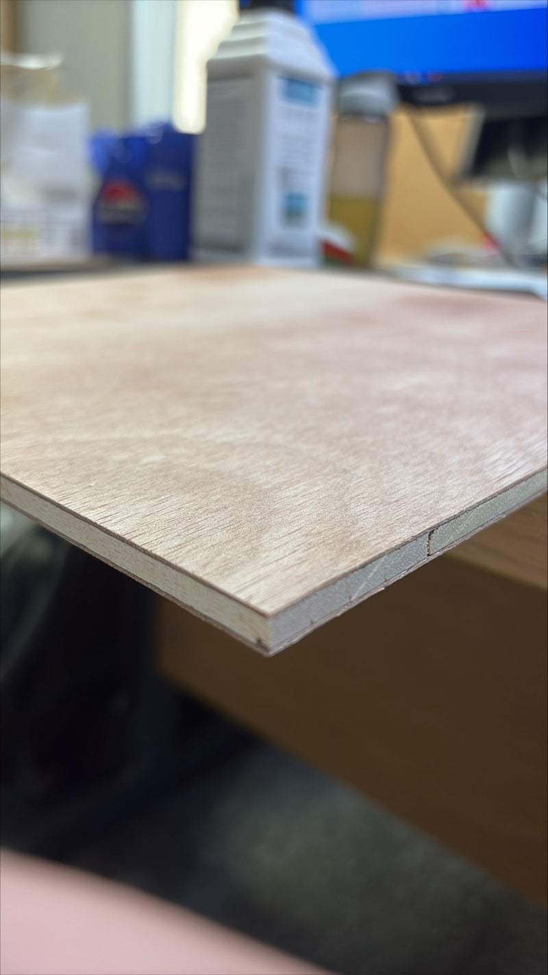 A close-up of a light-colored wood veneer sheet or panel, showing its grain texture and edge profile, photographed in a workshop or studio setting.