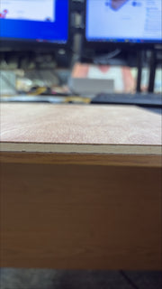 A close-up shot of a light brown wood veneer sheet with a visible grain texture and a clean edge, resting on a desk with blurred computer monitors in the background.