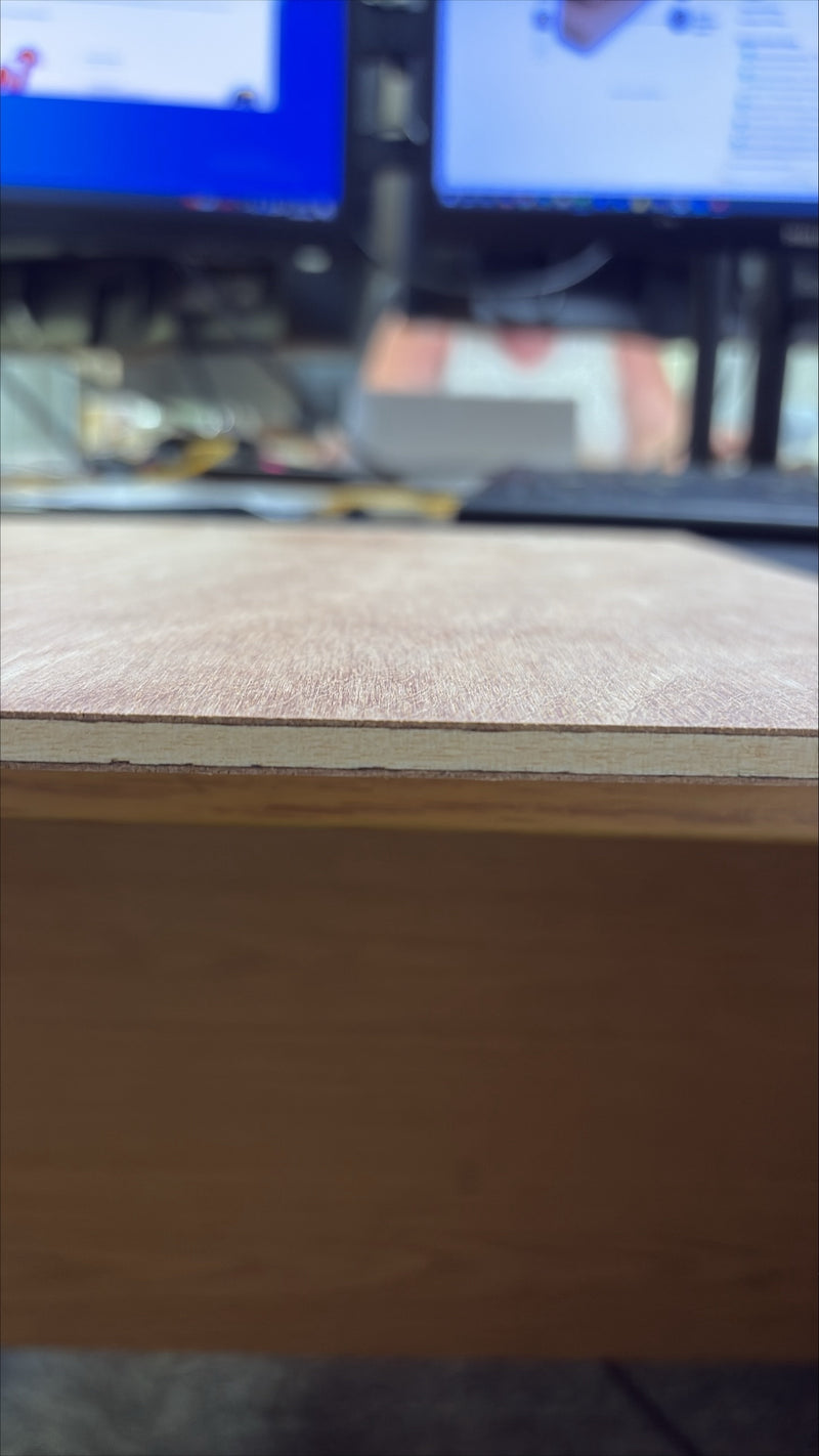 A close-up shot of a light brown wood veneer sheet with a visible grain texture and a clean edge, resting on a desk with blurred computer monitors in the background.