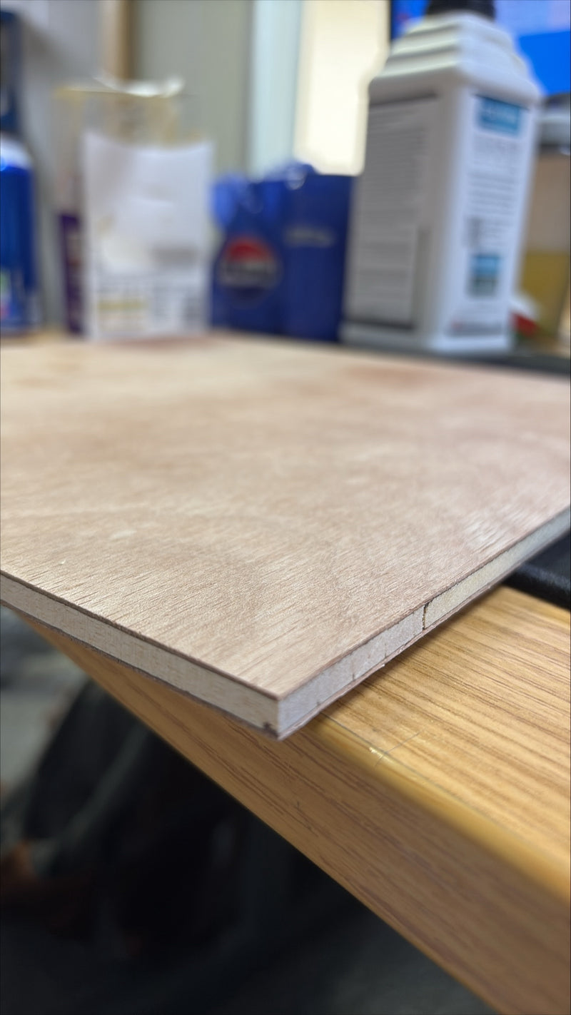 A close-up of a light wood laminate or veneer sheet with a visible edge, resting on a wooden table in what appears to be a workshop or craft space.