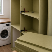 A set of olive green particle board shelves and panels shown installed inside a wooden cabinet, with a T-clamp holding one shelf in place. A white washing machine and a window are visible in the background, indicating a home or utility room environment.
