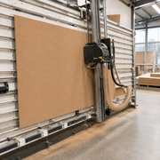 A large tan wood veneer sheet mounted on a vertical industrial CNC router or wood cutting machine inside a workshop. The machine features a black control box, a beige flexible dust collector hose, and a silver aluminum rail system.