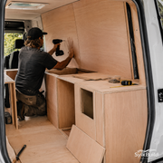 Person working on a wooden interior setup inside a van, using tools.